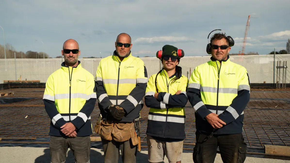 Four construction workers wearing fluorescent yellow and navy high-visibility safety jackets with reflective strips standing together on a construction site.